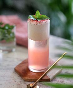 a tall glass filled with pink liquid and topped with a mint sprig next to two gold spoons