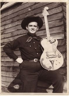 a black and white photo of a man in cowboy attire holding an electric guitar with his hands on his hips