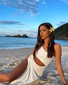 a beautiful woman sitting on top of a sandy beach next to the ocean with a flower in her hair