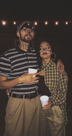 a man and woman standing next to each other in front of string lights holding coffee cups