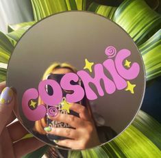 a person holding up a mirror with the word cosmic on it in front of some plants