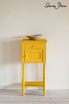 a small yellow table with a bowl on it's top sitting against a white wall