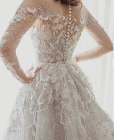 a woman wearing a wedding dress with long sleeves and flowers on the skirt, standing in front of a window