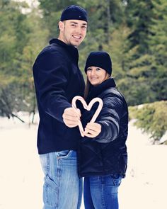 a man and woman standing in the snow holding up a heart shaped sign with trees in the background