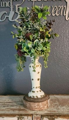 a white vase filled with lots of green plants on top of a wooden table next to a wall