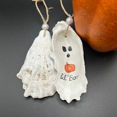 two ceramic ghost ornaments are hanging from twine strings next to an orange on the table