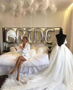 a woman sitting on top of a bed next to a wedding dress and balloon arch