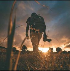 a man with a backpack and camera walking through tall grass in the sun setting behind him