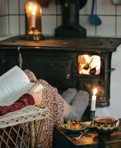 a woman sitting in a chair reading a book next to an open fire place with food on the table