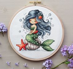 a cross stitch mermaid sitting on top of a table next to purple flowers