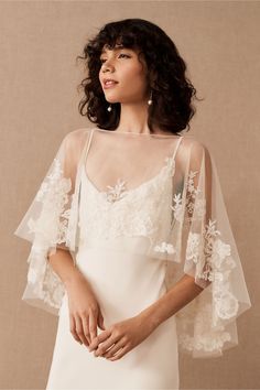 a woman wearing a white dress with sheer sleeves and flowers on the shoulder, standing in front of a beige background