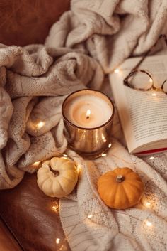 an open book and some candles on a table with blankets, pumpkins and glasses