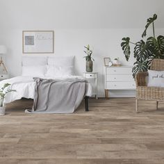 a bedroom with white walls and wood floors, a wicker chair, potted plants on the bed