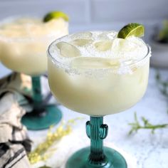 two margaritas sitting next to each other on top of a white countertop with lime wedges