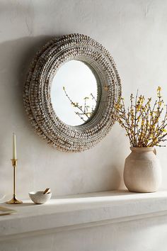 a white mantle topped with a vase filled with flowers and a round mirror hanging on the wall