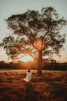 a woman in a white dress standing next to a tree on top of a field