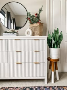 a white dresser sitting next to a mirror and potted plant