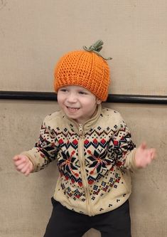 a young boy wearing an orange knitted hat and jacket, standing in front of a wall