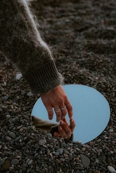 a person holding their hand in front of a circular mirror on the ground with gravel