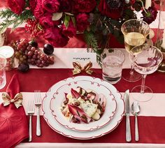 a table set with plates, silverware and wine glasses on it in front of red flowers