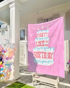 a birthday cake on a pink banner in front of a white house with balloons around it