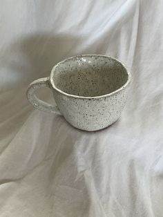 a white cup with speckles on it is sitting on a white cloth covered surface