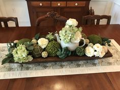 an arrangement of flowers and succulents in a white pitcher on a wooden table