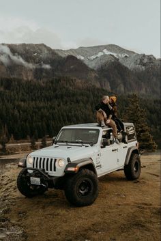 two people sitting on the top of a white jeep with mountains in the back ground