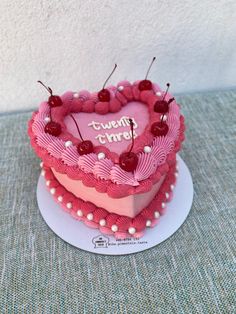 a pink heart shaped cake with cherries on top sitting on a table next to a white wall