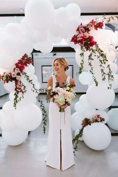 a woman in a long white dress holding a bouquet and standing next to some balloons