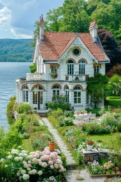 a white house sitting on top of a lush green hillside next to a lake with lots of flowers