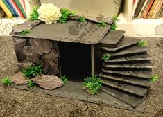 a small house made out of stone with plants and rocks on the outside, in front of a bookshelf