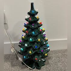 a green ceramic christmas tree with multi - colored lights on it's base, next to a white wall