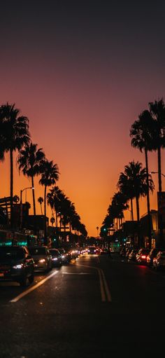 cars are driving down the street at night with palm trees in the foreground and an orange sky