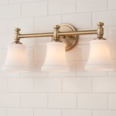 three light bathroom fixture with white glass shades on the bottom and brass finish, against a white brick wall