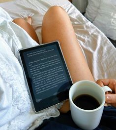 a woman laying in bed with a cup of coffee and an electronic book on her lap