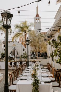 a long table set up with white linens and greenery for an outdoor event