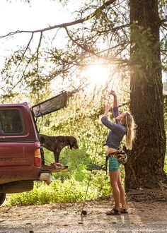 a woman standing next to a dog in the back of a pick up truck on a dirt road