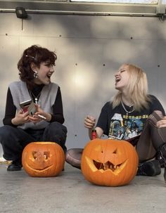 two women sitting on the ground with pumpkins in front of them and one is holding a cell phone