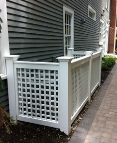 a white trellis fence next to a gray house on a brick sidewalk in front of a building