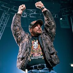 a man in camo jacket and hat raising his hands up at an outdoor concert