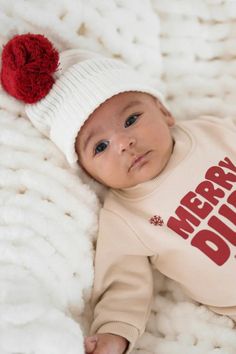 A baby lying on a soft, white, textured blanket, wearing a beige outfit with the words "Merry Dude" in bold red letters and a white knit hat with a large red pom-pom. Pompom Beanie, White Beanie, White Beanies, Snow Days, Cozy Accessories, Cotton Beanie, Kids Beanies, Cold Weather Outfits