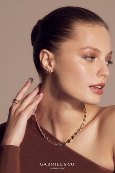 a woman wearing a brown top and gold jewelry with her hand on her shoulder, looking at the camera