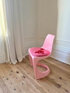 a pink chair sitting on top of a hard wood floor next to a window with curtains