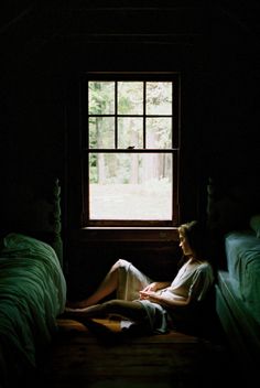 a woman sitting in front of a window with her legs crossed on the bed and looking out