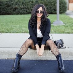 a woman sitting on the curb with her legs crossed, wearing black stockings and boots