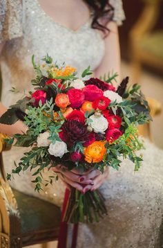 a bride holding a bouquet of red and orange flowers in her hand while sitting on a chair