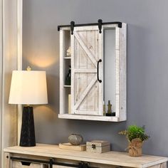 a white cabinet sitting next to a lamp on top of a wooden table in front of a gray wall