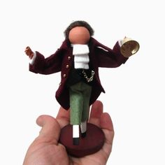 a hand holding a small toy doll in it's right hand, wearing a red jacket and green pants