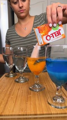 a woman is pouring blue liquid into three glasses on a wooden table with orange and blue liquids in them